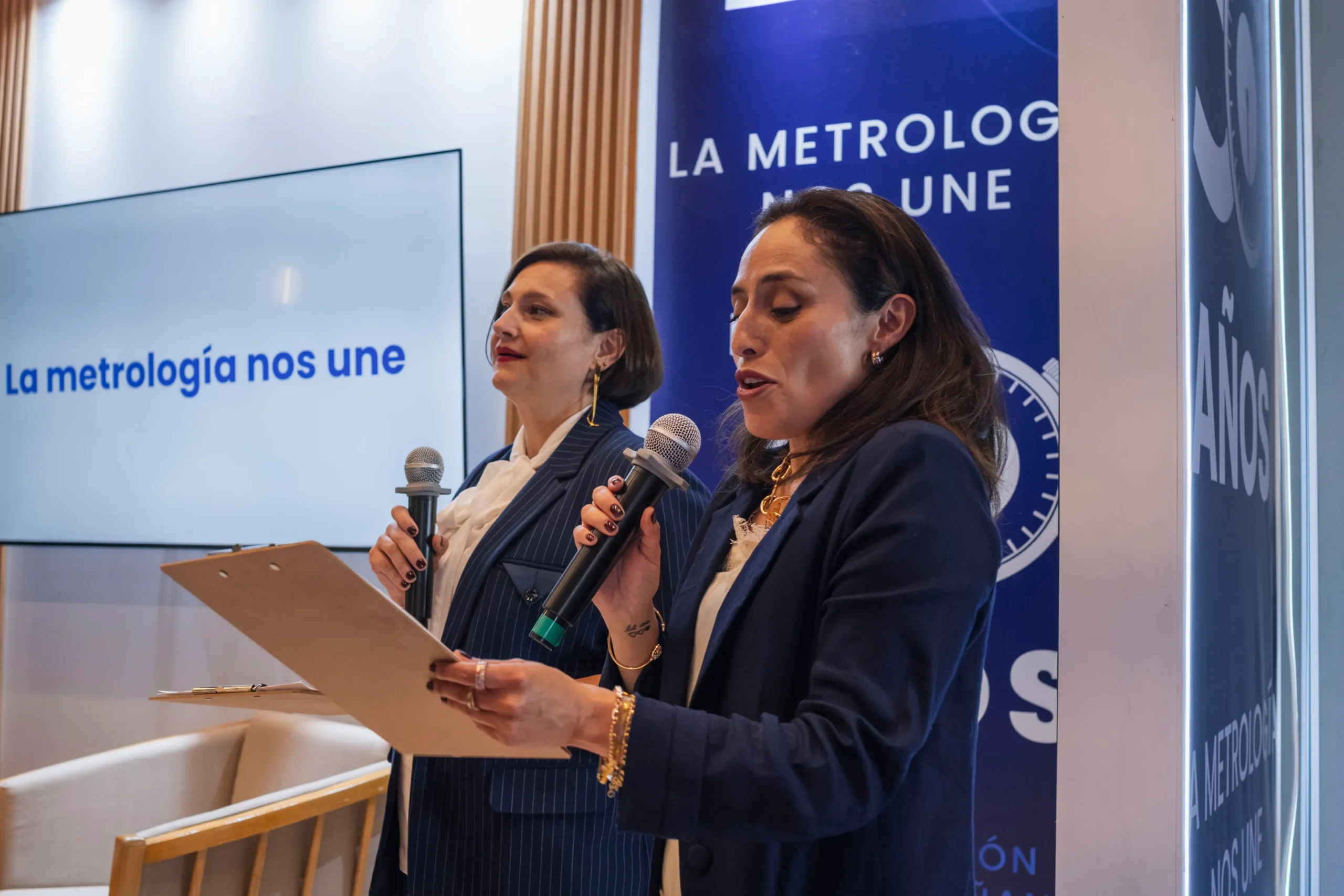 Dos mujeres conferencistas hablando por micrófono durante un evento de metrología con una pantalla de fondo que dice La metrología nos une